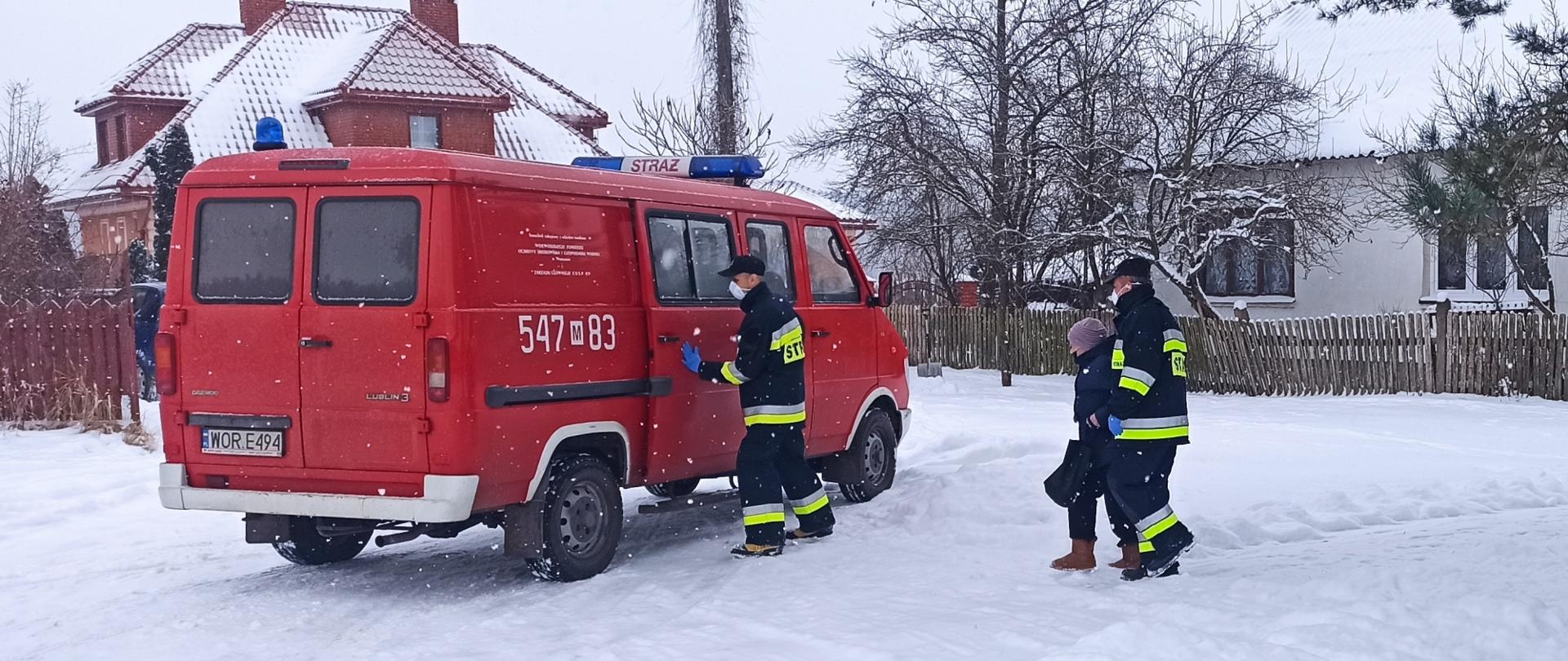 Strażacy wspierają Narodowy Program Szczepień. W centrum zdjęcia samochód strażacki, bus, czerwony. Dwóch strażaków w mundurze bojowym pomaga wsiąść do pojazdu starszej kobiecie. Samochód stoi na zaśnieżonej ulicy. W tle budynki mieszkalne.