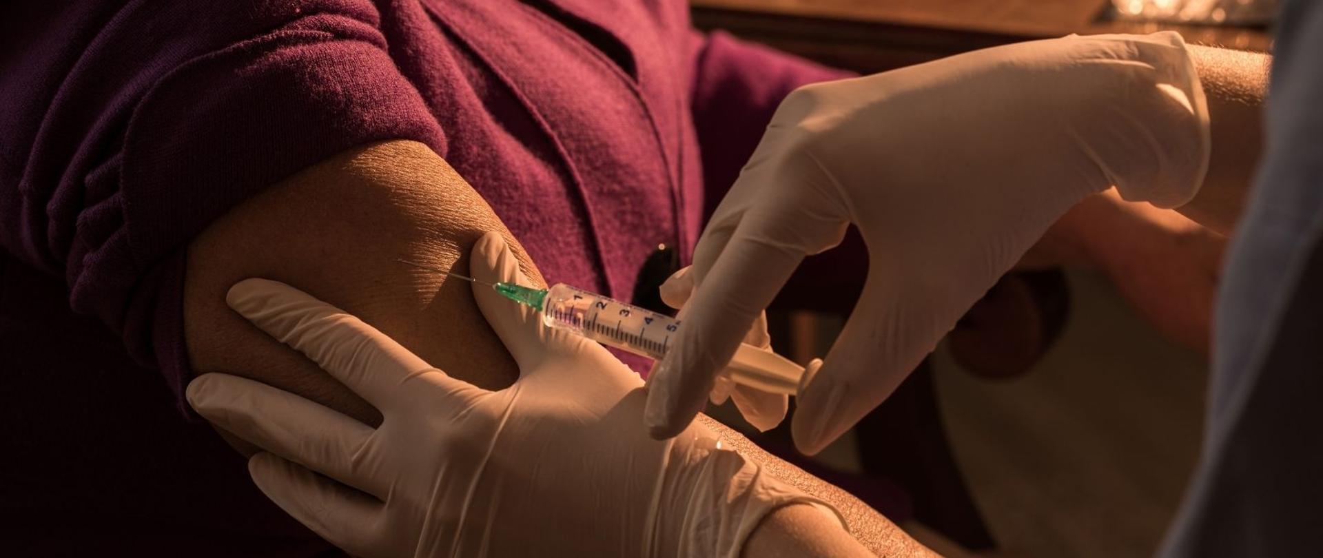 Podanie szczepionki w ramię starszej osoby. Na zdjęciu widoczna bordowa bluzka pacjentki i jej ramię oraz dwie ręce pielęgniarki w białych rękawiczkach, która strzykawką wstrzykuje szczepionkę