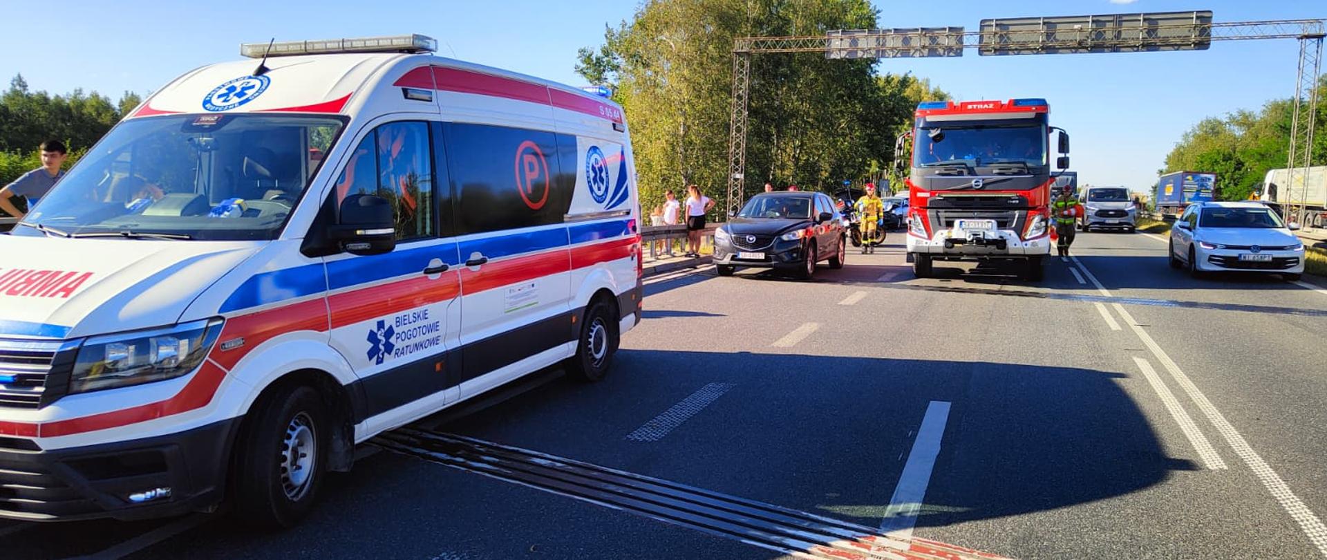 Zdjęcie przedstawia wypadek drogowy, a na miejscu są już obecne służby ratunkowe, w tym karetka Pogotowia Ratunkowego i wóz strażacki, oraz grupka osób na poboczu.
