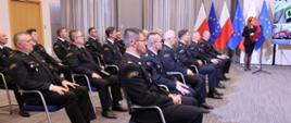 Funkcjonariusze Państwowej Straży Pożarnej ubrani w mundury wyjściowe siedzą na krzesłach obok stoi kobieta za nią stoją flagi Unii Europejskiej, Polski. Przed kobietą stoi mikrofon. 