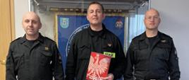 Trzech mężczyzn w mundurach Państwowej Straży Pożarnej stoi przodem ustawieni do wspólnego zdjęcia, za nimi widoczna jest gablota w której wisi sztandar Komendy powiatowej PSP w Pleszewie