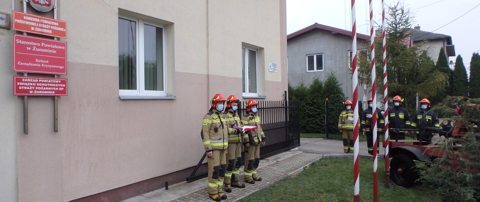 Trójka strażaków stojąca w żółtych ubraniach specjalnych, w czerwonych hełmach na tle budynku Państwowej Straży Pożarnej w Żurominie. Strażak stojący po środku trzyma na rękach flagę Polski. Pozostali stoją w pozycji zasadniczej. Po prawej stronie stoi czworo strażaków stojących w pozycji zasadniczej na tle drzew.