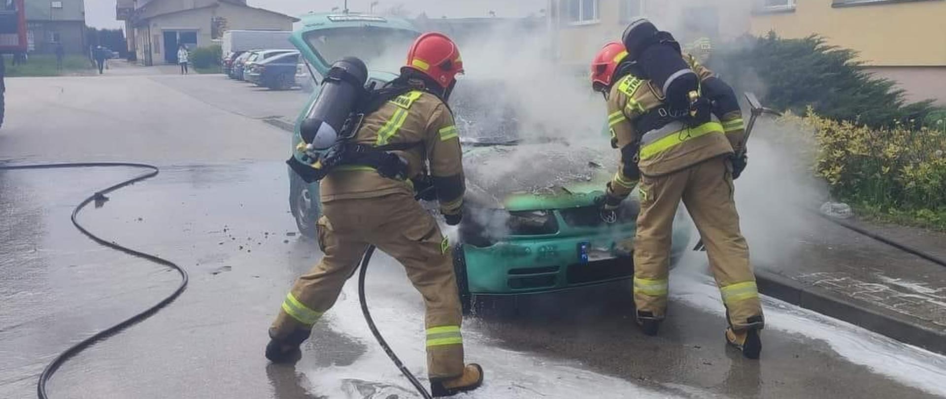 Dwóch strażaków w ubraniu specjalnym oraz sprzęcie ochrony układu oddechowego gasi pożar samochodu osobowego. Z prądownicy podawana jest piana ciężka.