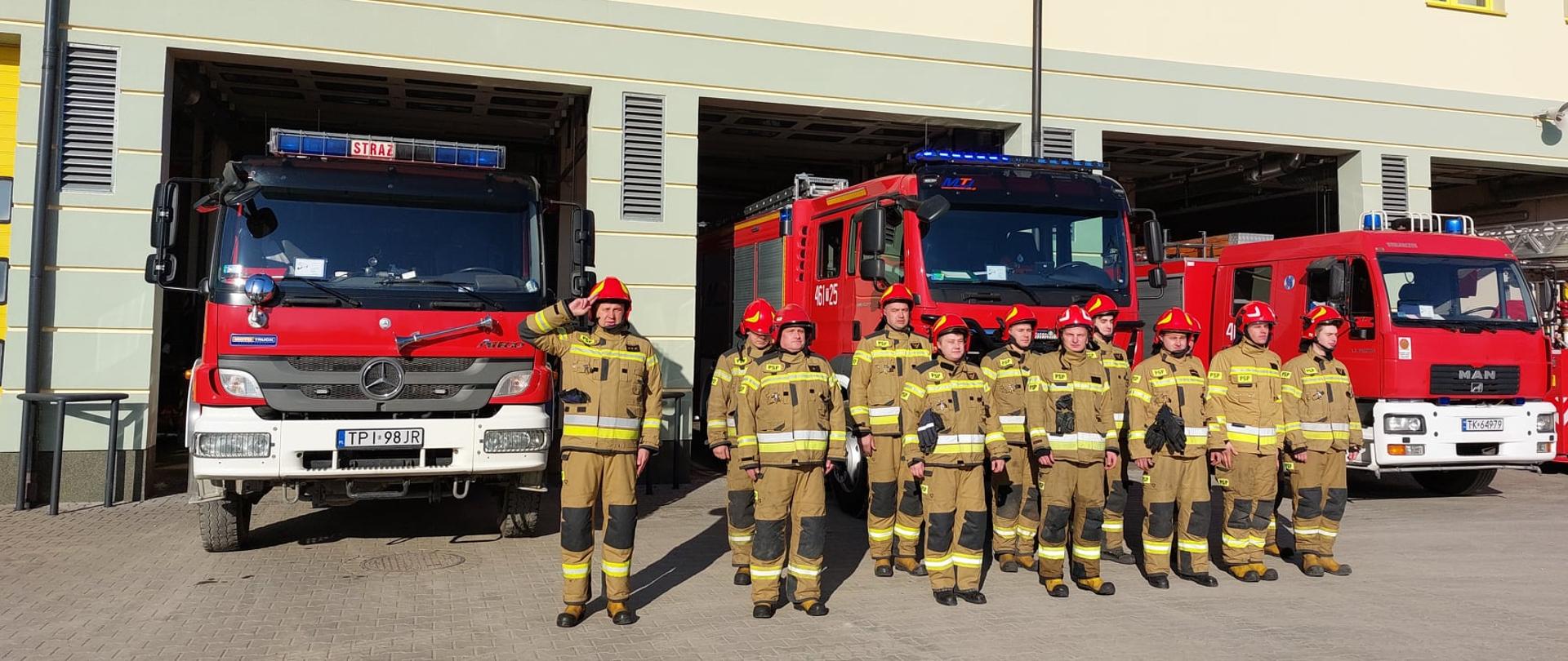 Zdjęcie przedstawia strażaków w ubraniach specjalnych oddających hołd poległym strażakom z Ukrainy, w tle pojazdy strażackie z włączonymi sygnałami świetlnymi.