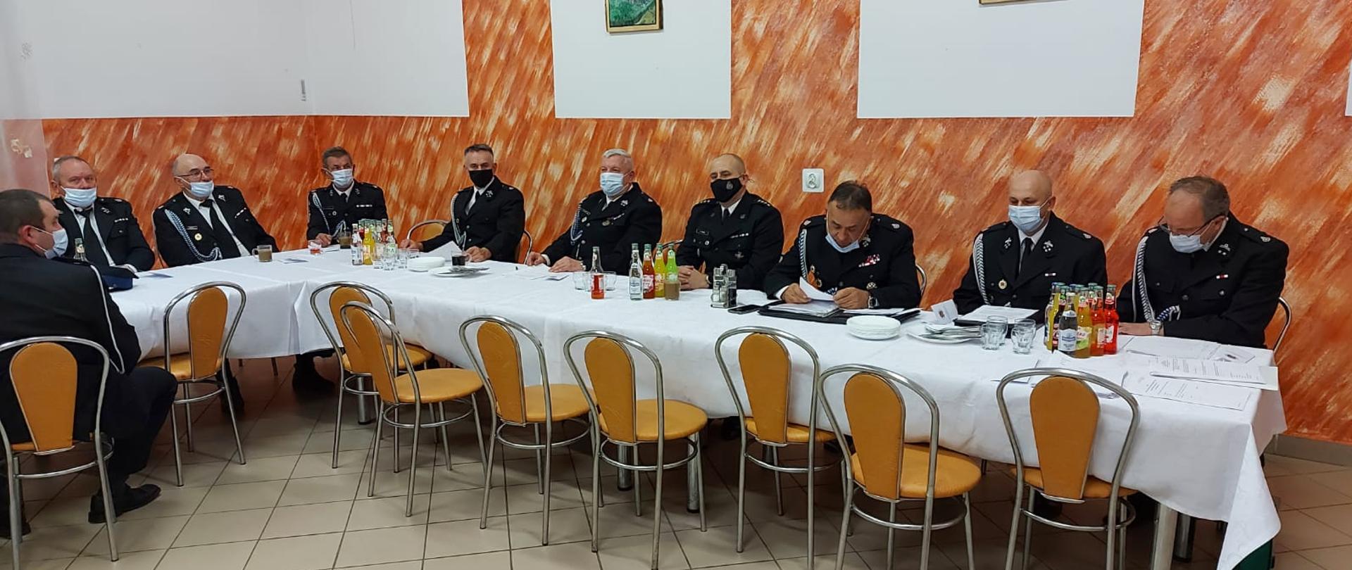 Na zdjęciu widać strażaków OSP oraz komendanta powiatowego PSP w Ustrzykach Dolnych w mundurach galowych którzy siedzą przy stołach na zebraniu OSP. Na stołach są białe obrusy oraz napoje. 