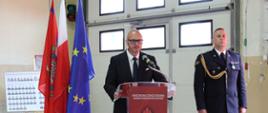 W centralnym punkcie, za czerwoną mównicą z logo Państwowej Straży Pożarnej, stoi mężczyzna w ciemnym garniturze i okularach, który przemawia do mikrofonu. Po jego prawej stronie, w postawie zasadniczej, stoi strażak w ciemnym mundurze galowym ze sznurem funkcyjnym i odznaczeniami na piersi.
W tle widoczna jest duża, biała brama garażowa z małymi okienkami, a obok niej pali się zielone światło sygnalizatora. Po lewej stronie mównicy ustawiony jest poczet sztandarowy z trzema flagami: strażacką, polską oraz unijną. Na ścianie w głębi widać tablicę ze zdjęciami personelu jednostki.