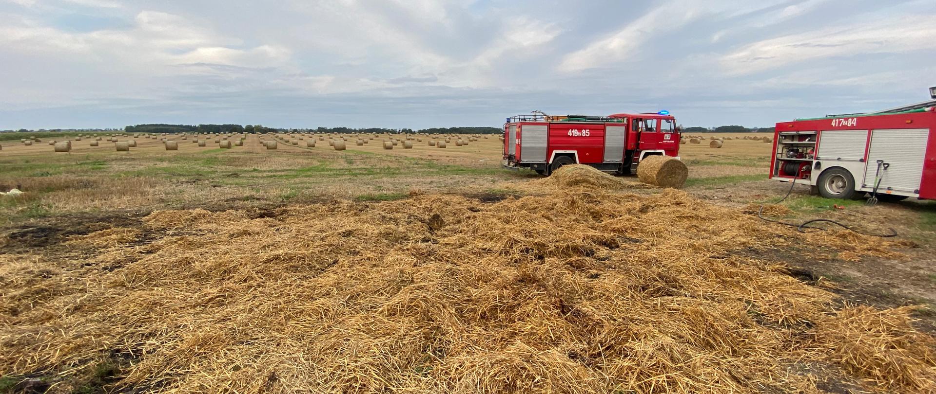 Na fotografii wykonanej na środku pola znajdują się dwa wozy strażackie - jeden w centrum, drugi po prawej stronie. Pomiędzy wozami na środku znajduje się rozgarnięta słoma. W oddali na polu znajdują się snopki, a na końcu pola zaczyna się las. Zdjęcie wykonano w dzień przy lekkim zachmurzeniu.