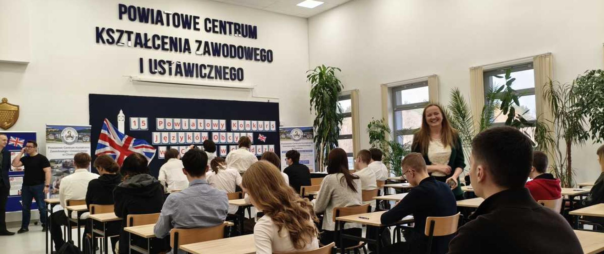 To zdjęcie przedstawia grupę uczniów uczestniczących w konkursie językowym w Powiatowym Centrum Kształcenia Zawodowego i Ustawicznego.
Wydarzenie: Zdjęcie dokumentuje 15. Powiatowy Konkurs Języków Obcych.
Miejsce: Wydarzenie odbywa się w sali lekcyjnej Powiatowego Centrum Kształcenia Zawodowego i Ustawicznego.
Uczestnicy: W sali znajdują się uczniowie siedzący przy ławkach oraz osoby pełniące prawdopodobnie rolę organizatorów lub nauczycieli.
Otoczenie: Sala jest wyposażona w ławki szkolne, a w tle widać dekoracje związane z konkursem, w tym flagę Wielkiej Brytanii.