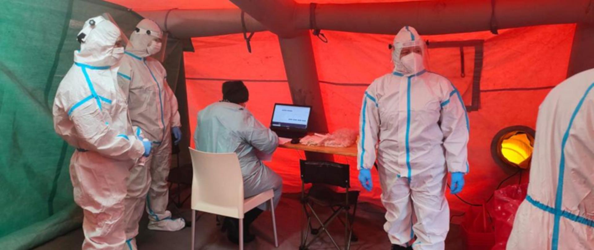 na zdjęciu, w czerwonym namiocie pięć osób ubranych w białe kombinezony ochronne, maseczki i przyłbice, cztery osoby stoją, jedna siedzi przy komputerze