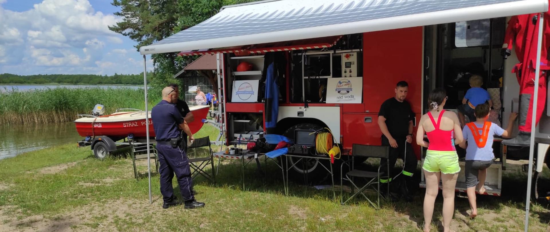 Zdjęcie przedstawia samochód strażacki, łódź, policjanta strażaka i młodzież. 