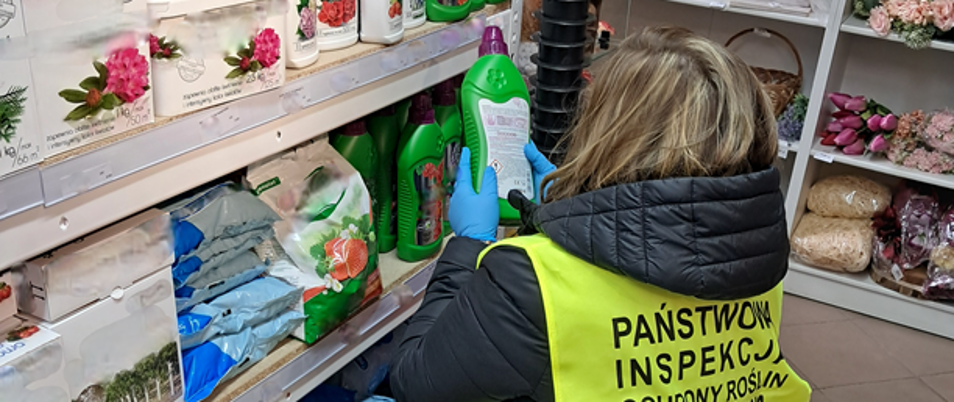 Inspektor znajdujący się w sklepie ubrany w żółtą, odblaskową kamizelkę czyta etykietę produktu, aby sprawdzić informacje w niej zawarte. W tle widać półki z produktami nawozowymi.