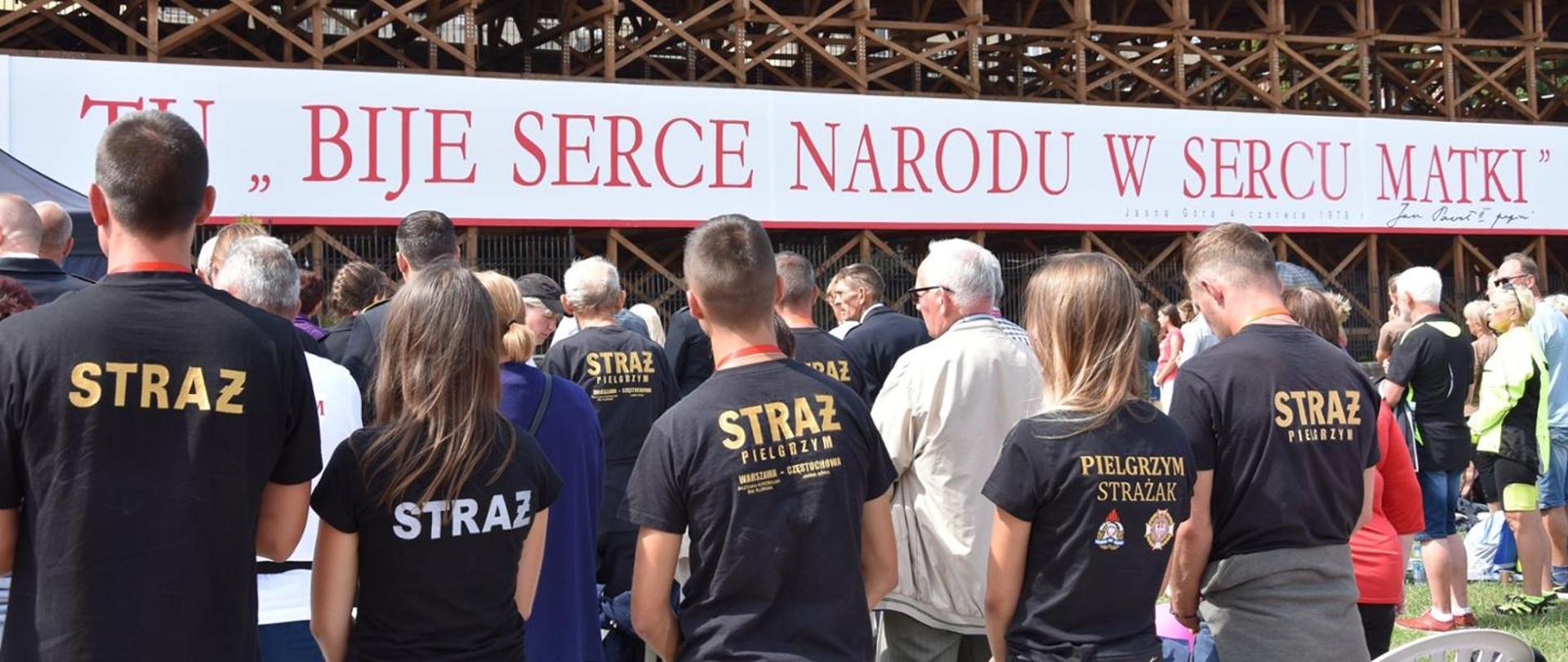 Zdjęcie przedstawia strażaków w czarnych koszulkach z napisami STRAŻ. Nad nimi czerwony napis na białym tle TU BIJE SERCE NARODU W SERCU MATKI