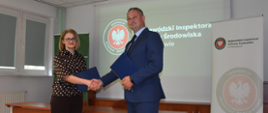Uścisk dłoni po podpisaniu porozumienia o współpracy pomiędzy Mazowieckim Wojewódzkim Inspektorem Ochrony Środowiska Krzysztofem Cząstkiewiczem i Mazowieckim Wojewódzkim Lekarzem Weterynarii Izabelą Floryszczyk. Przedstawiciele obu inspekcji stoją na sali konferencyjnej, przed stołem na którym podpisali porozumienie.