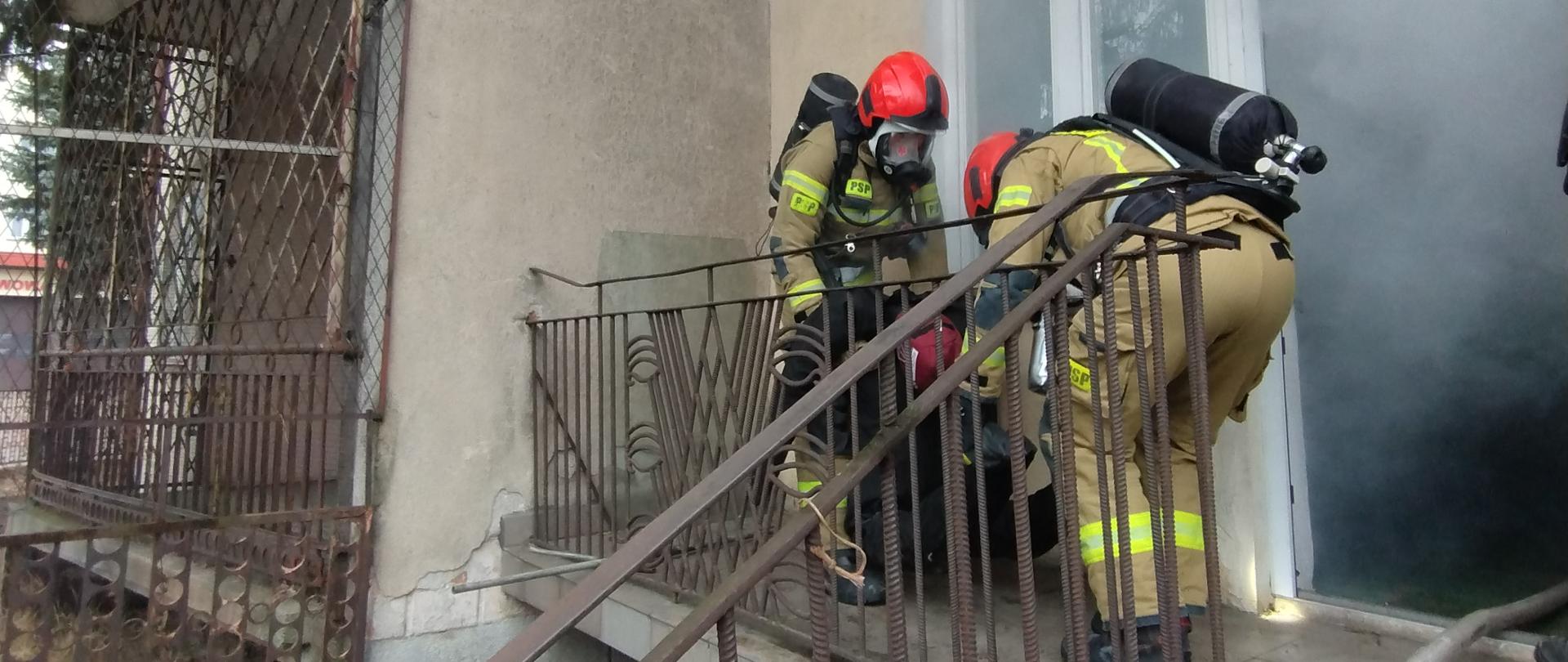 Strażacy w ubraniach specjalnych, aparatach powietrznych i z linią gaśniczą wynoszą manekina z zadymionego domu.