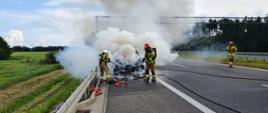 Pożar auta na S5. Jezdnia trasy S5 w kierunku Poznania. Na pasie awaryjnym stoi palący się samochód osobowy. Wokół niego silne zadymienie. Przed nim w kłębach dymu dwóch strażaków gaszących pożar. Na pasie ruch strażak kierujący akcją prowadzi rozmowę przez radiotelefon. W tle las i łąka.