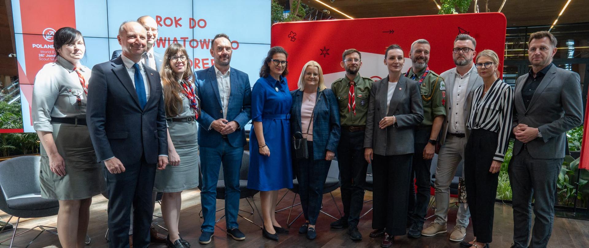 Grupowe zdjęcie jedenastu osób, ubranych w stroje formalne i harcerskie mundury, stojących na tle ekranu z napisem „ROK DO ŚWIATOWEGO JAMBOREE”.