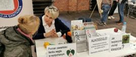 Kobieta obsługująca stoisko PSSE rozmawia z uczestniczką wydarzenia, na stole wyeksponowane stojaki wypełnione ulotkami.
