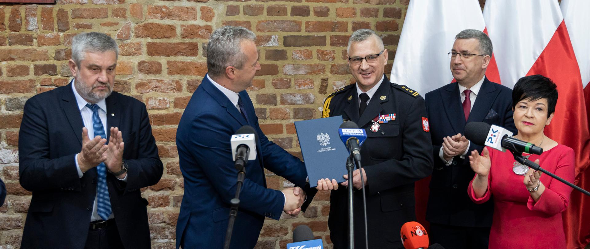 Trwa konferencja prasowa. Za stołem prezydialnym stoją włodarze regionu i miasta, parlamentarzyści i kujawsko-pomorski komendant wojewódzki PSP. W tle fragment starego muru, należącego do murów obronnych miasta, na nim godło i logo PSP, a za plecami gości flagi narodowe. Przed rozmówcami mikrofony. Wojewoda wręcza dokument komendantowi wojewódzkim.