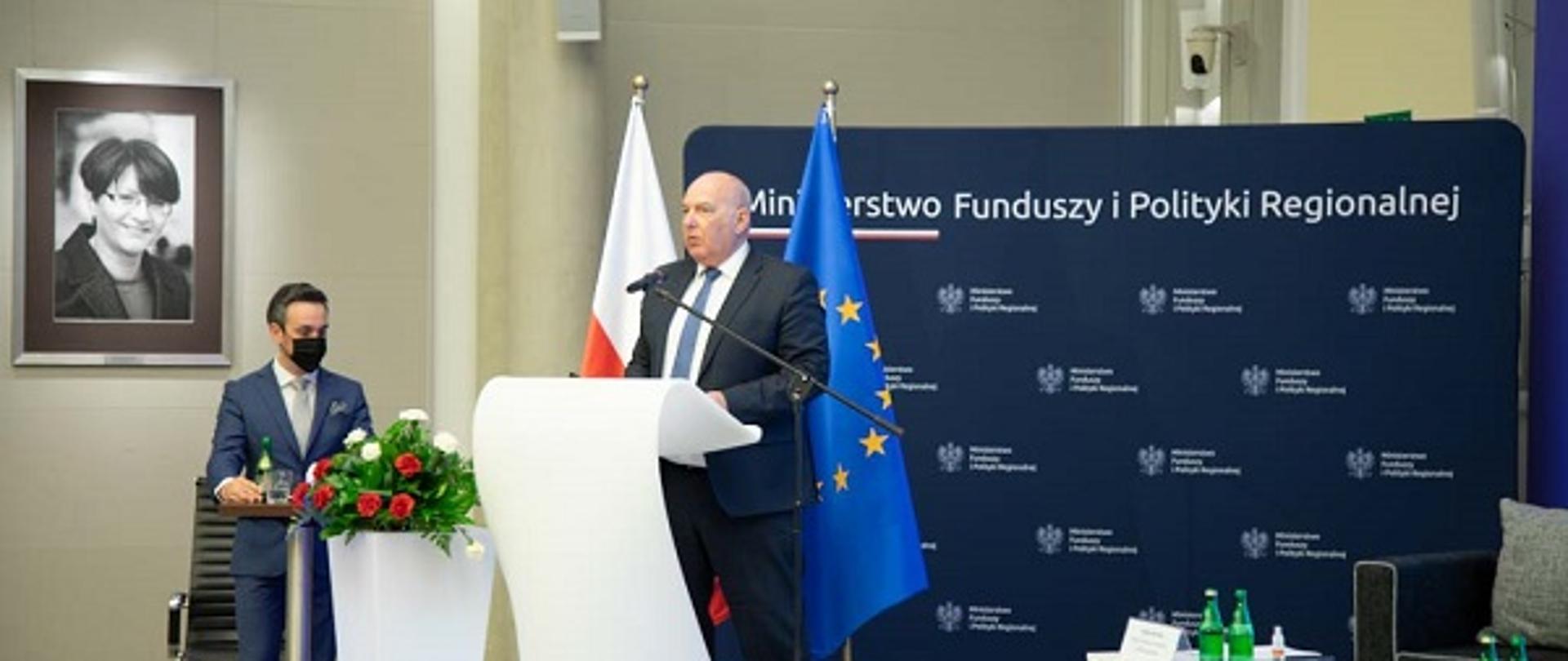 Minister Tadeusz Kościński przemawia na scenie. W tle ścianka z logo Ministerstwa Funduszy i Polityki Regionalnej