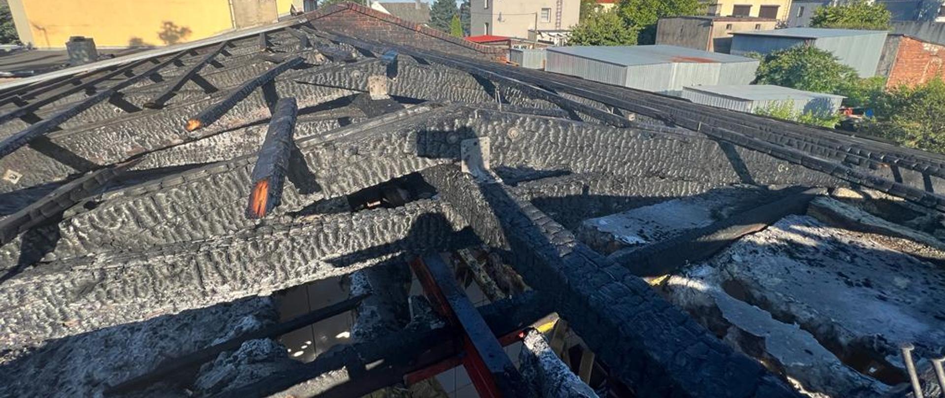 Widok z dachu budynku. Czarne spalone drewniane krokwie na całym dachu. W tle inne budynki 