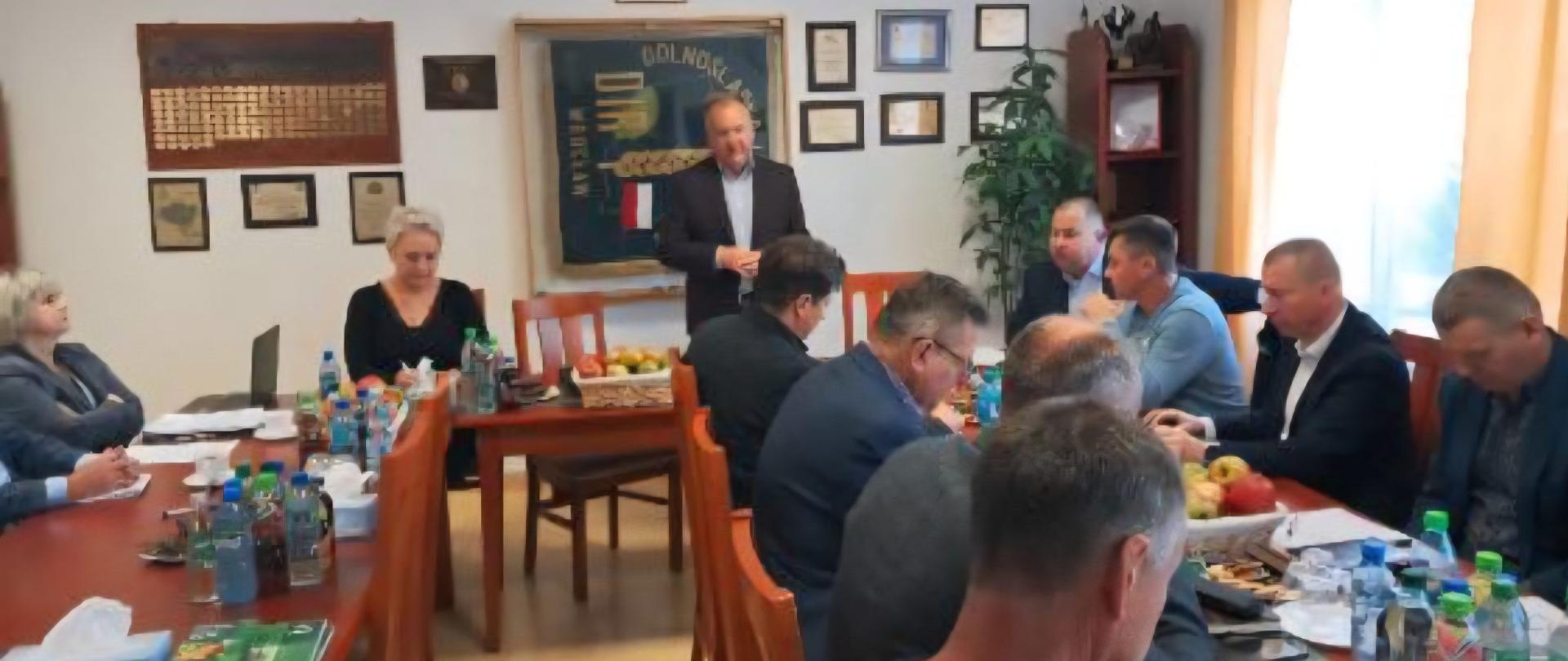 Uczestnicy posiedzenia siedzący przy stołach w gabinecie. Na stolach leżą owoce, napoje oraz liczne dokumenty.