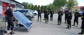 Po prawej stronie zdjęcia widać 10 strażaków. Po przeciwnej stronie stoi skierowany do nich twarzą instruktor prezentujący trenażer fotowoltaiczny. Dwa panele fotowoltaiczne nazywane trenażerem znajdują się na platformie jezdnej tuż obok instruktora.
Wszyscy strażacy są w czarnych ubraniach koszarowych z czapkami z daszkiem na głowach.
Plac na którym odbywają się zajęcia jest pokryty asfaltem, po lewej stronie w tle znajduje się szary budynek z dwiema czerwonym bramami garażowymi przed którym stoją samochody osobowe.
