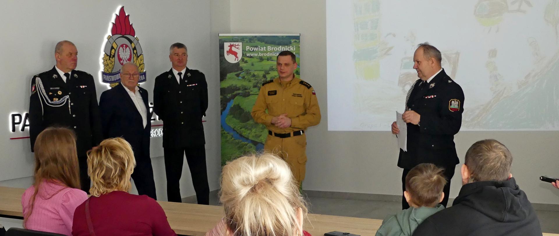 Zdjęcie przedstawia moment powitania laureatów eliminacji powiatowych Ogólnopolskiego Strażackiego Konkursu Plastycznego – Komendant Powiatowy Straży w Brodnicy wita zgromadzone dzieci wraz z opiekunami. Goście siedzą w ławkach na pierwszym planie. W tle obok Komendanta stoją przedstawiciele organizatora konkursu. Na ścianie wyświetlana jest praca jednego z uczestników. Za Komendantem stoi baner Powiatu Brodnickiego a na nim zielone krajobrazy i wijąca się środkiem rzeka.