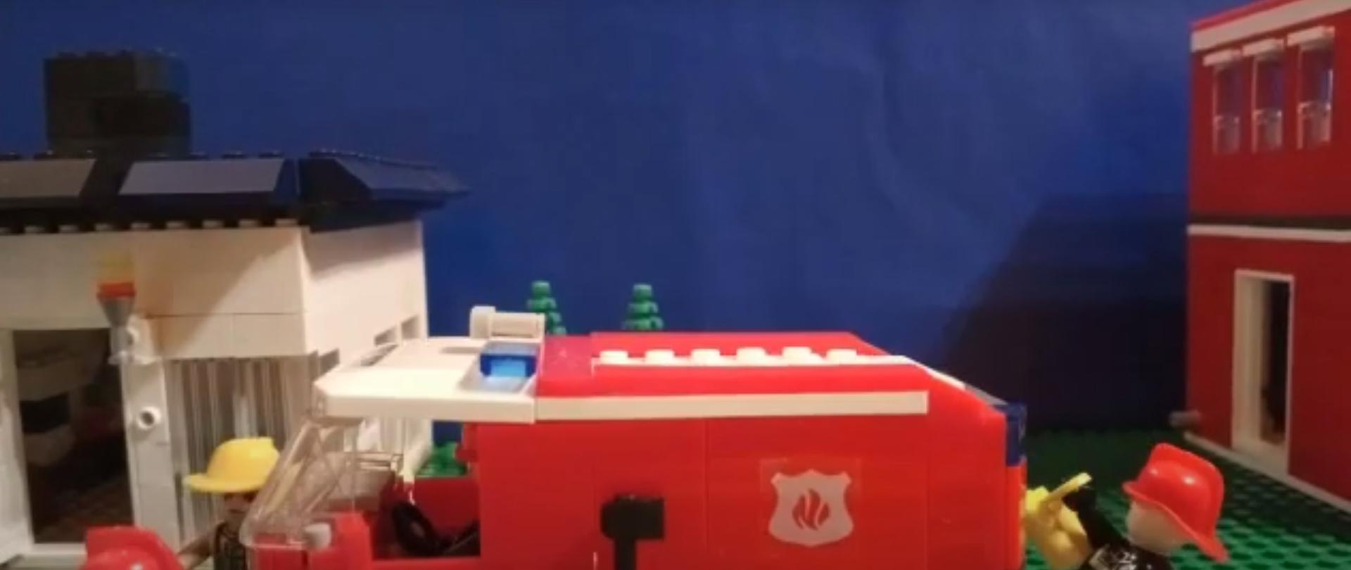 Zdjęcie przedstawia zrobionych z klocków LEGO strażaków w akcji. Na środku znajduje się czerwona straż, obok niej stoi dwóch strażaków. W tle widać dom oraz w prawym krańcu siedzibę PSP.