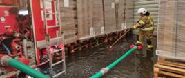 Usuwanie skutków intensywnych opadów deszczu. Zdjęcie przedstawia wnętrze zalanego przez wodę magazynu opakowń tekturowych. Wewnątrz znajduje się wóz pożarniczy, do którego podłączony jest zielony wąż. Strażak nogą przytrzymuje czerwony smok ssawny. W rękach trzyma natomiast miotłę.
