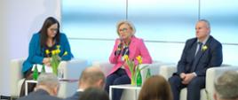 Przy małych białych stolikach siedzi wiceminister Machałek w różowej marynarce, kobieta w niebieskiej marynarce i mężczyzna w garniturze, wiceminister mówi do trzymanego w ręku mikrofonu, na stolikach stoją żółte tulipany w wazonach, przed stolikami na krzesłach siedzą ludzie.