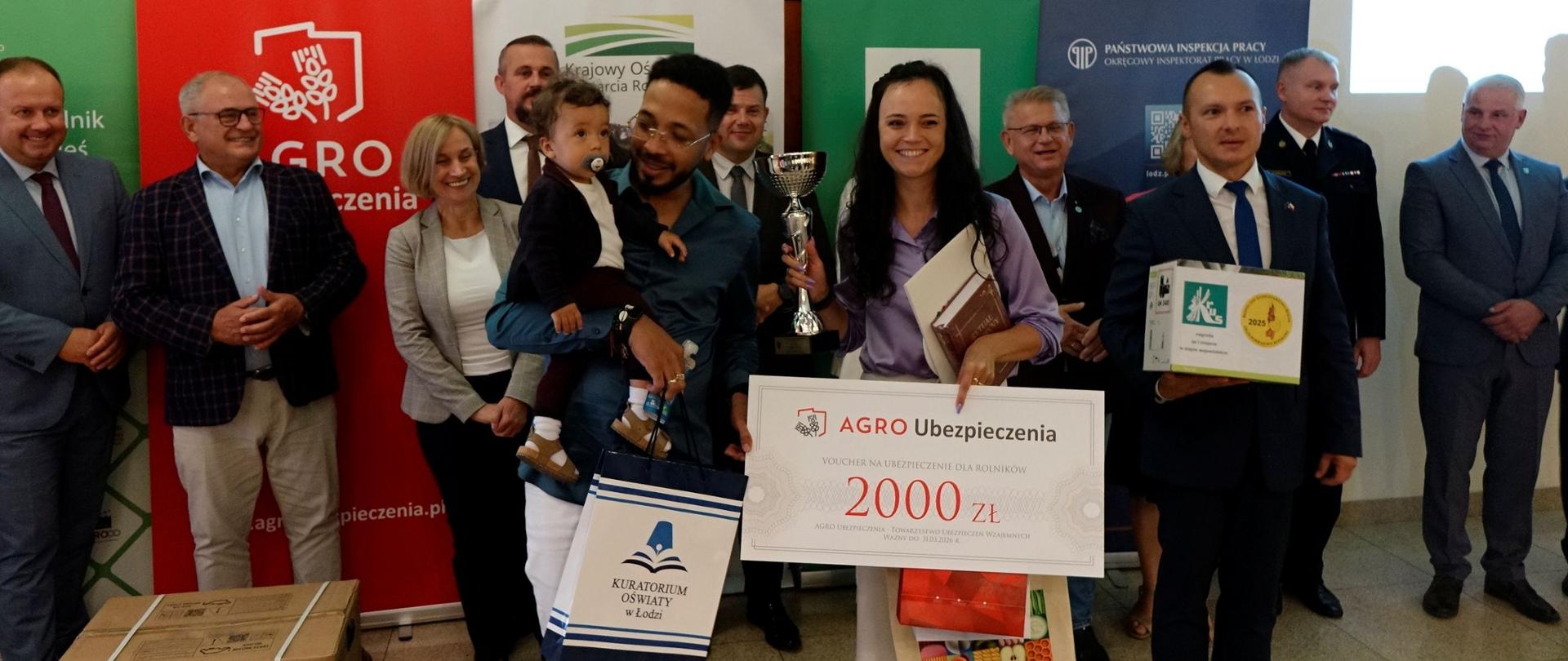 Na dużej sali stoi młoda uśmiechnięta kobieta ze srebrnym pucharem, symbolicznym czekiem na 2000 zł. i innymi nagrodami w rękach. Obok stoi mężczyzna z małym dzieckiem na ręku. Po lewej stronie stoją dwa pudła z nagrodami. Z tyłu za nimi stoi grupa przedstawicieli organizatorów, z tyłu rollupy organizatorów.