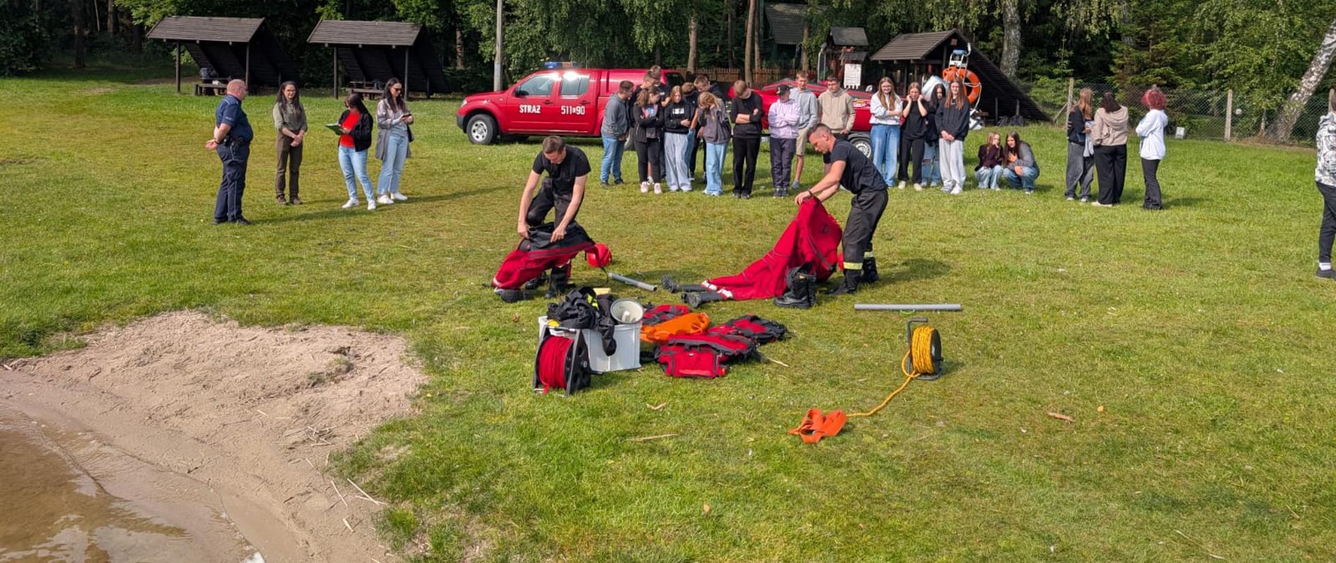 Dwóch strażaków na trawiastej plaży nad jeziorem ubiera się w skafandry suche do pracy w wodzie. W tle młodzież oraz strażacki pojazd nissan navara.