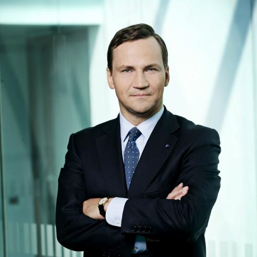 Radosław Sikorski, kolorowy portret stojącego mężczyzny, ciemne włosy, ciemny niebieski krawat w kropki, biała koszula, czarna marynarka, ręce założone na piersiach, na lewej dłoni zegarek, w tle szklane ściany.