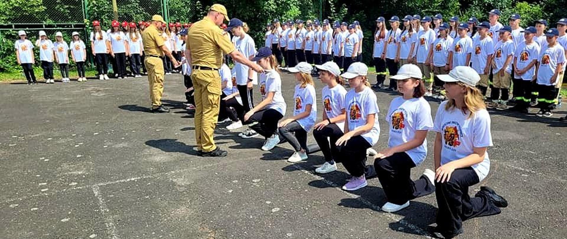 Zdjęcie zrobione na zewnątrz podczas uroczystej zbiórki uczestników obozu Młodzieżowych Drużyn Pożarniczych. Uczestnicy obozu, członkowie Młodzieżowych Drużyn Pożarniczych składają ślubowanie.
