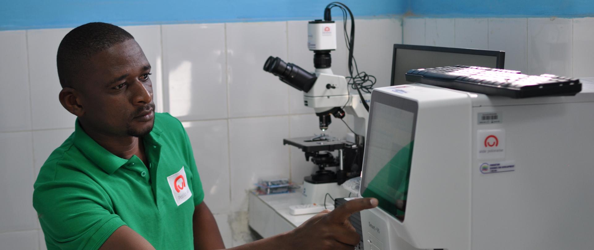 Mężczyzna w zielonej koszulce polo z logo polskiej pomocy obsługuje nowoczesną aparaturę laboratoryjną