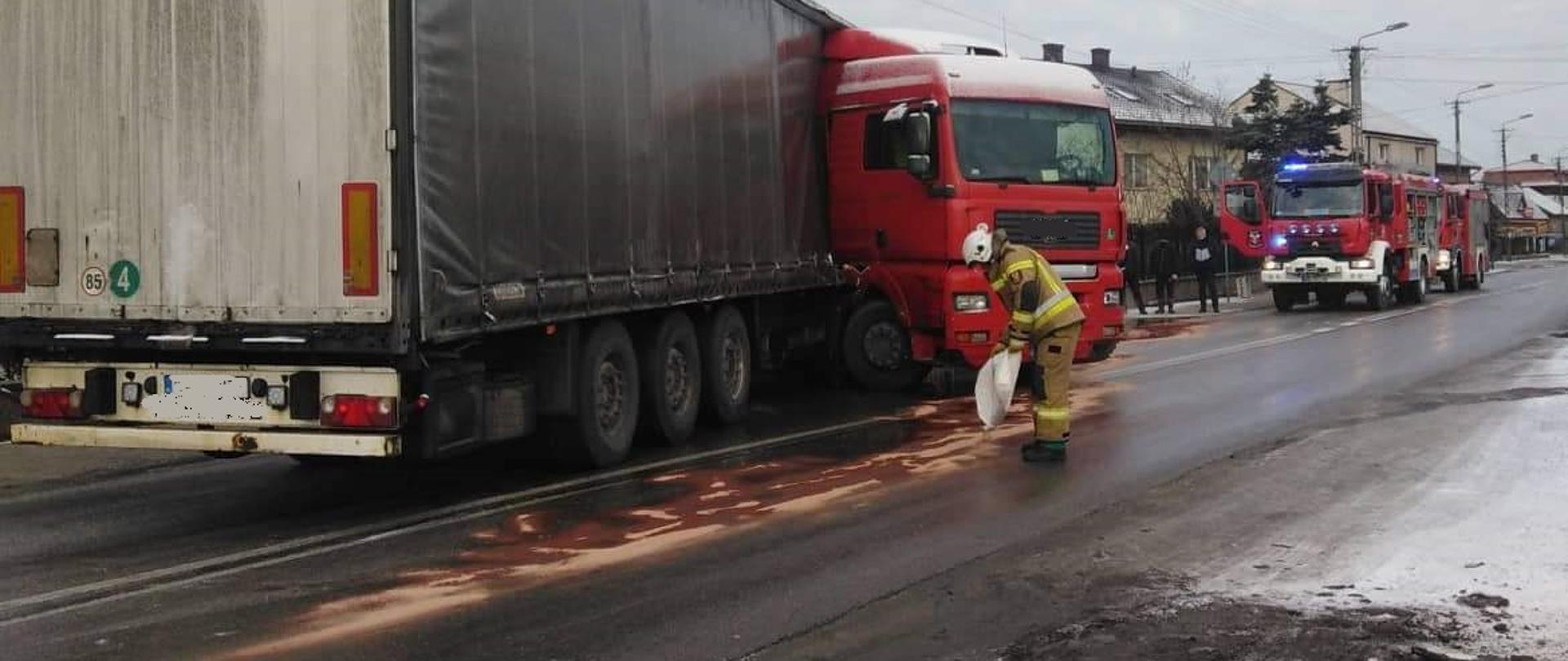 Na zdjęciu widać samochód ciężarowy z naczepą wysoką w kolorze szarym, na naczepie znajdują się znaki ostrzegawcze prostokątne w kolorze pomarańczowym ciągnik siodłowy w kolorze czerwonym znajduje się pod kątem prostym w stosunku do naczepy. Samochód znalazł się w tej pozycji ze względu na niekontrolowany poślizg pojazdu na łuku drogi, wskutek czego rozerwał się zbiornik z paliwem, z którego na jezdnię wyciekło ok. 800 litrów oleju napędowego. Na zdjęciu znajduje się strażak posypujący jezdnię sorbentem wokół pojazdu, który wchłania substancję. U góry nad samochodem znajduje się linia energetyczna, słupy oświetleniowe, oraz wystający czubek drzewa świerk, z prawej strony zdjęcia w oddali znajdują się trzy osoby postronne, kilka budynków mieszkalnych oraz dwa samochody ratowniczo-gaśnicze w kolorze czerwonym, poza obrysem jezdni znajduje się cienka warstwa śniegu, jezdnia ślizga z powodu ujemnej temperatury.