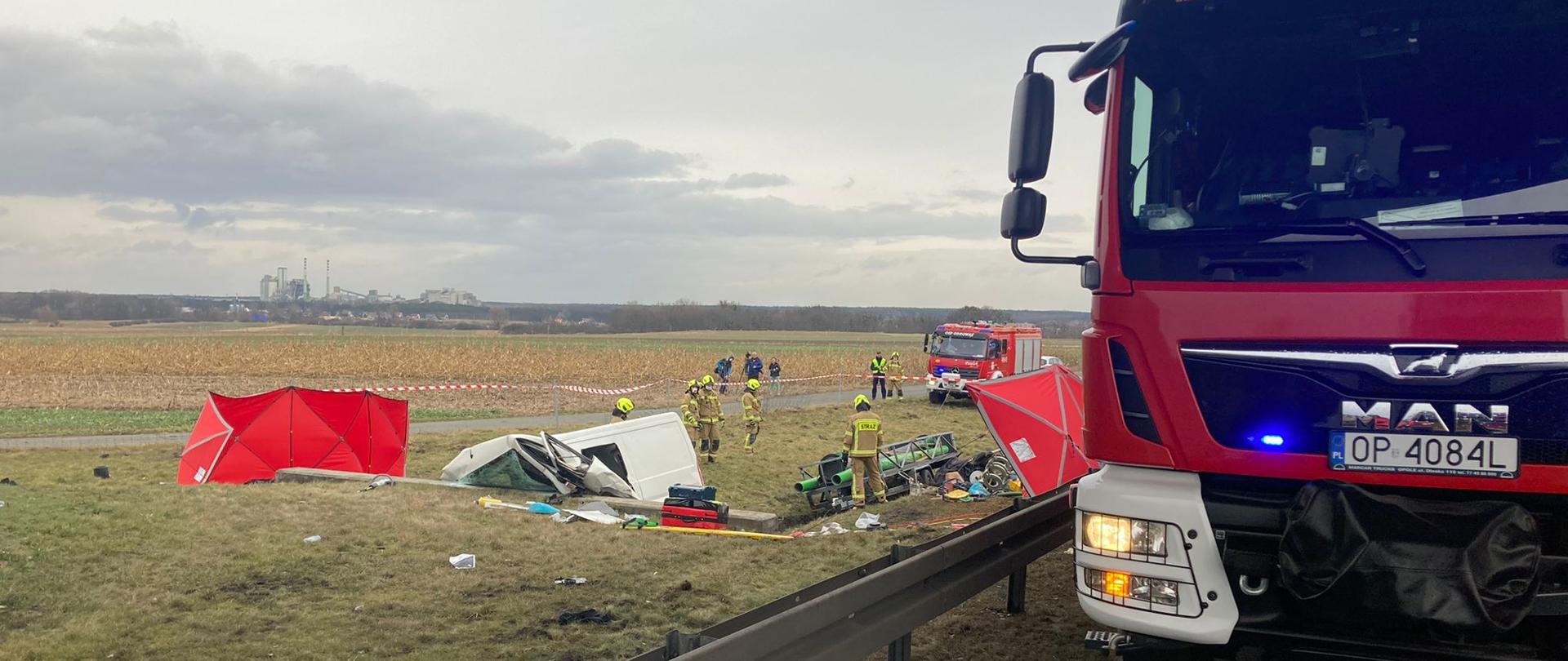 Strażacy PSP i OSP widoczni z oddali usuwają skutki wypadku drogowego samochodu dostawczego koloru białego jadącego z przyczepą. Działania trwają na poboczu w pobliżu przepustu melioracyjnego. Na zdjęciu widoczny przód samochodu pożarniczego stojącego na pasie awaryjnym autostrady tuż przy barierach energochłonnych.