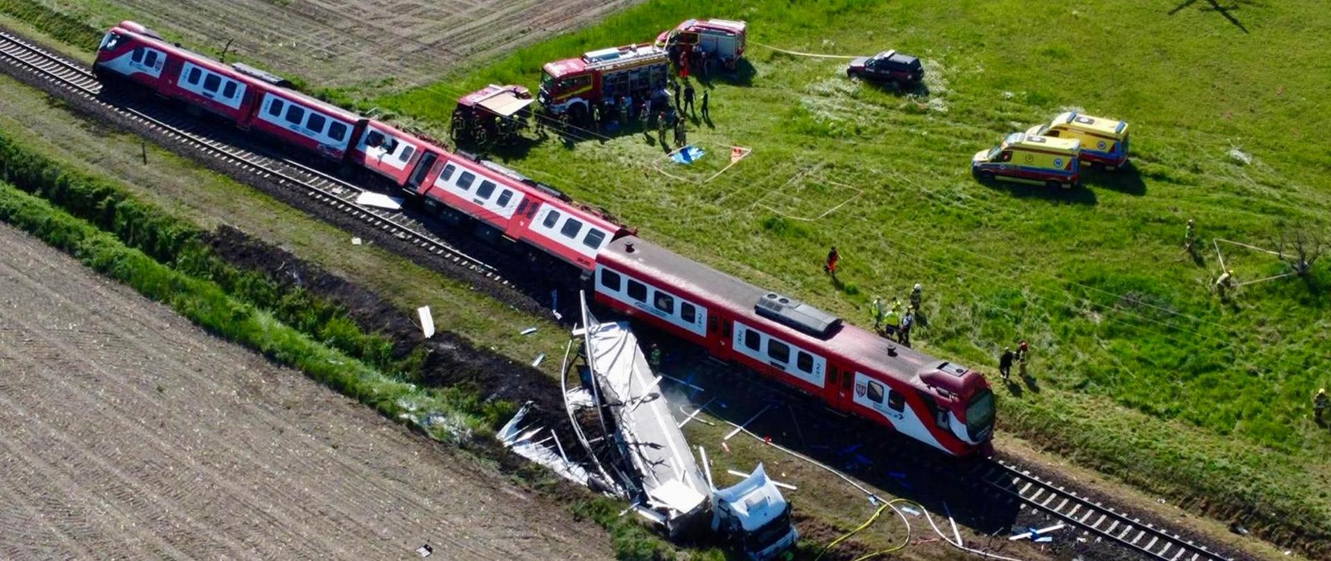 Widok z lotu ptaka na miejsce wypadku kolejowego na terenach wiejskich. Czerwono-biały pociąg pasażerski stoi na torach po zderzeniu z ciężarówką przewożącą ładunek ponadgabarytowy. Wokół widoczne są służby ratunkowe, wozy strażackie, karetki oraz śmigłowiec ratunkowy