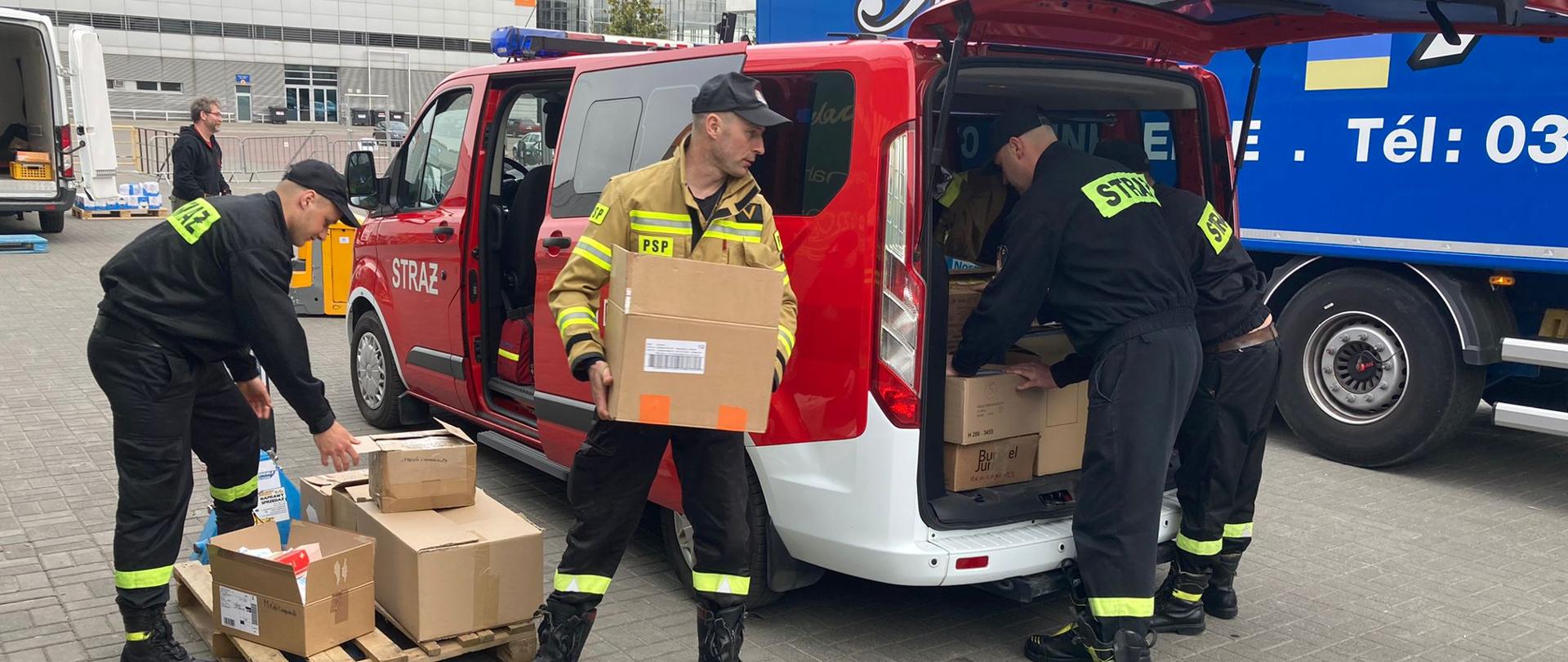 Na zdjęciu strażacy załadowują kartony do czerwonego samochodu strażackiego