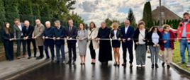 Grupa oficjeli stoi w poprzek drogi, trzymając wstęgę. Uczestnicy mają w dłoniach nożyczki i symbolicznie przecinają wstęgę na znak otwarcia nowej drogi.