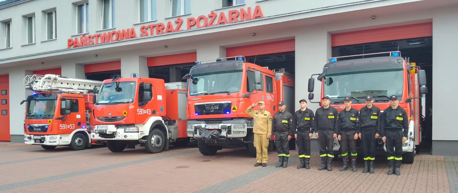 Strażacy przed samochodami strażackimi na tle komendy PSP