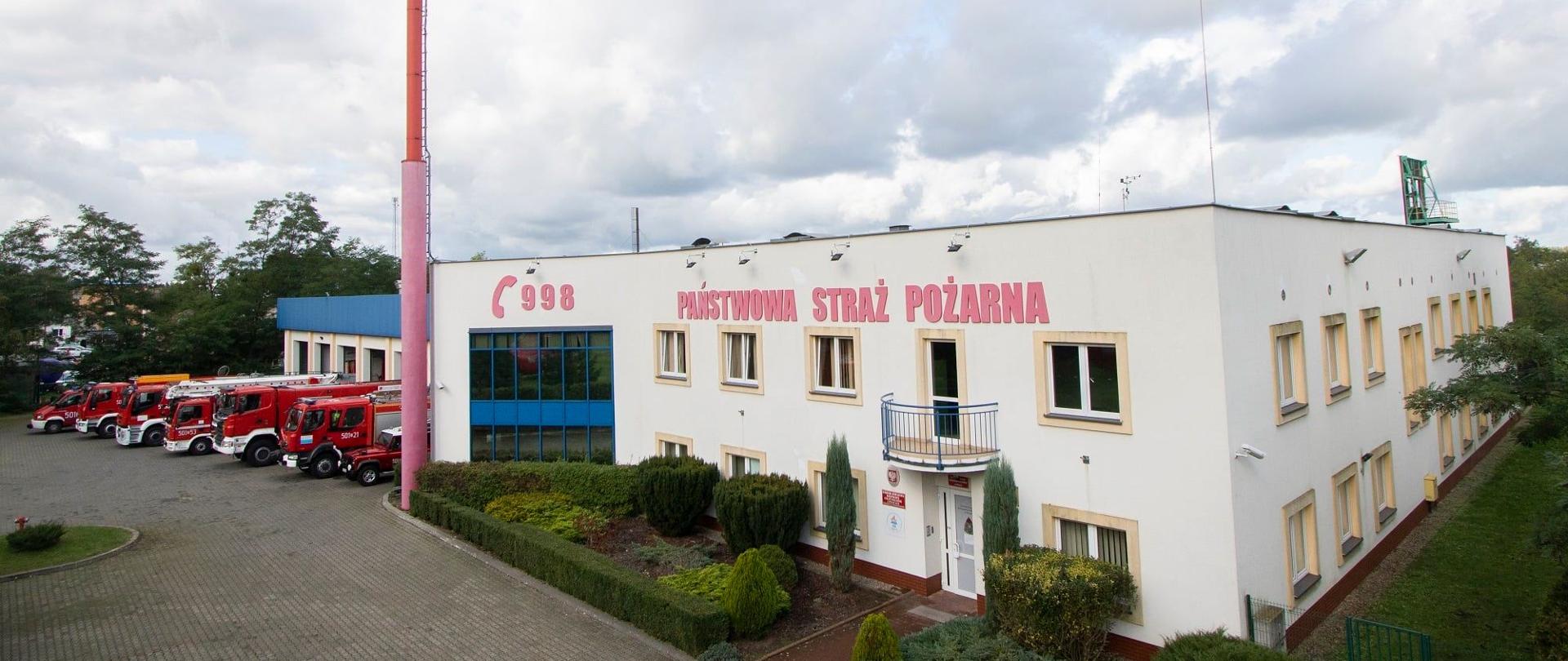 Biały budynek z napisem Państwowa Straż Pożarna. Na rogu budynku czerwony maszt. Przed bramami stoją samochody pożarnicze