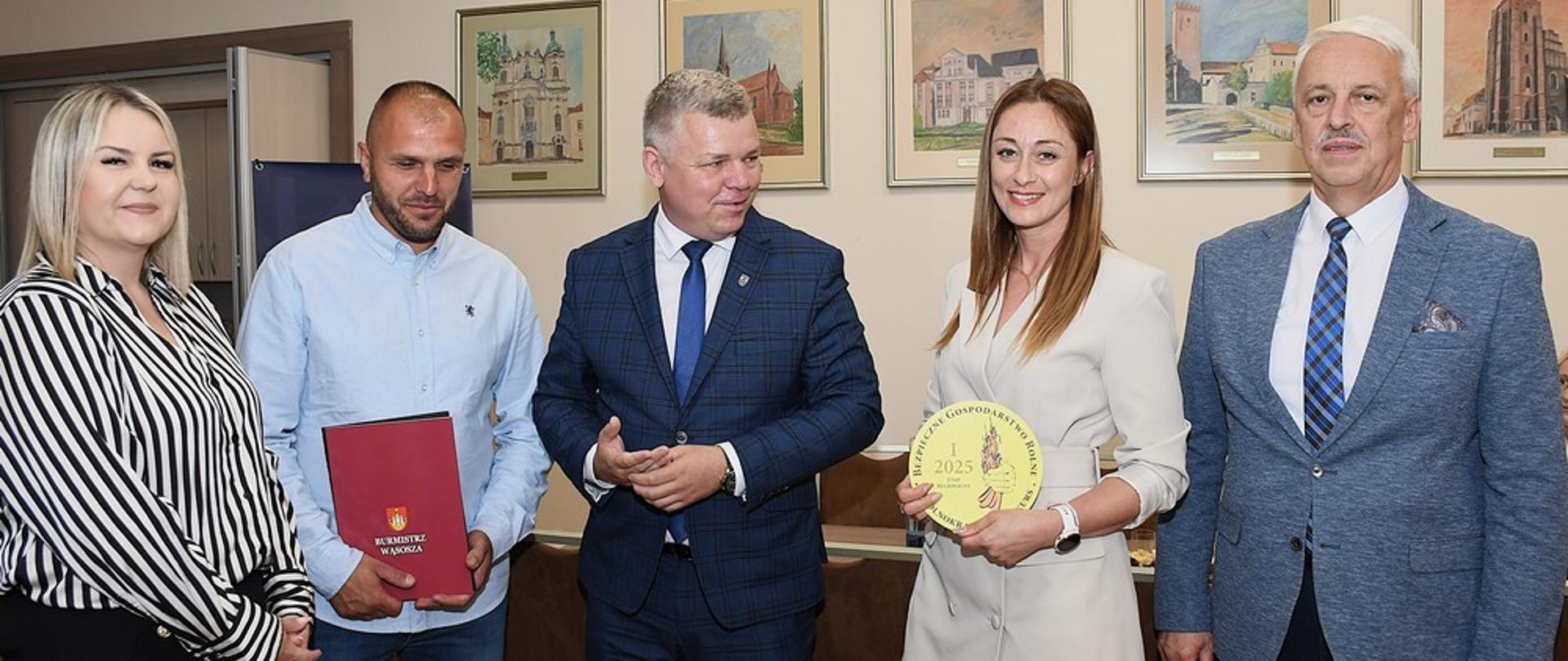 W sali na tle grafik wiszących na ścianach stoją dwie kobiety i trzech mężczyzn. Jedna z kobiet trzyma w rękach okrągłe logo konkursu BGR, a jeden z mężczyzn po lewej trzyma w rękach czerwoną ozdobną teczkę z dyplomem. Wszyscy uśmiechają się do zdjęcia.