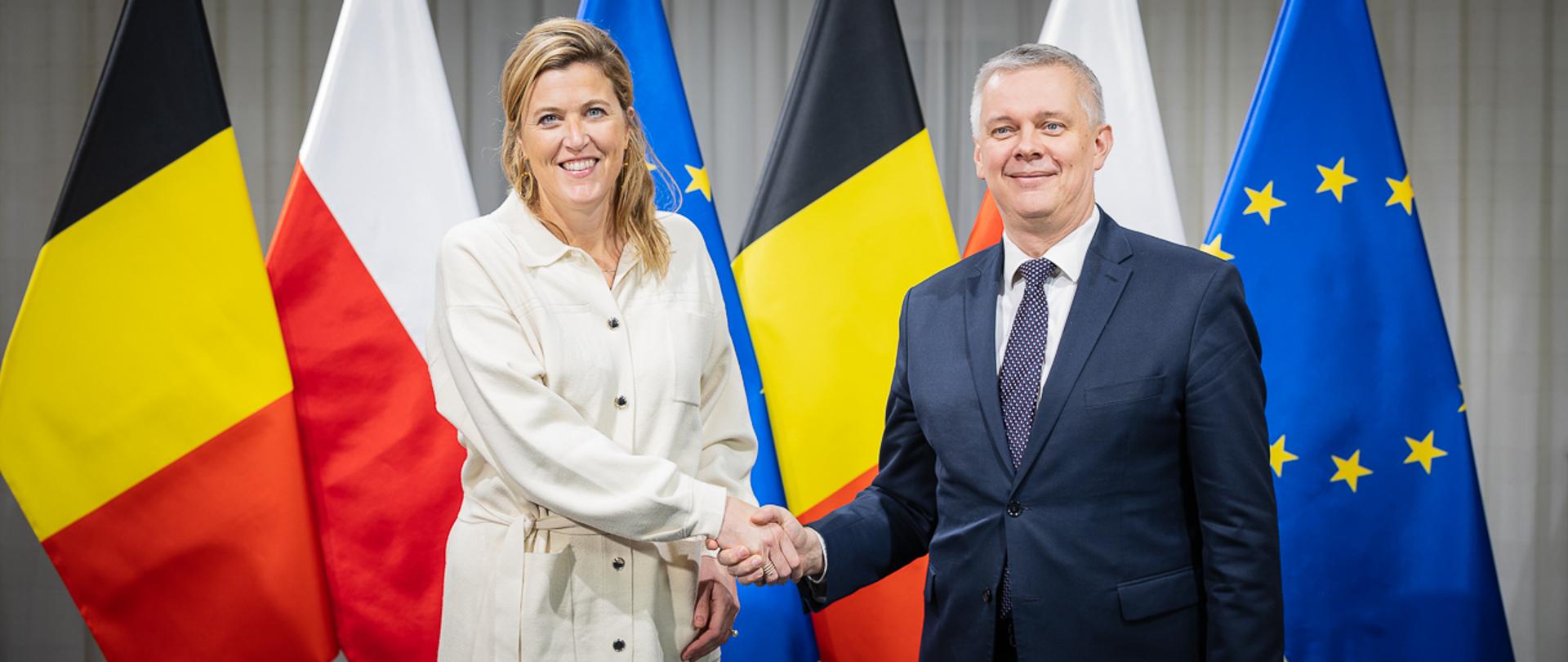 Kobieta i mężczyzna w geście powitania na tle flag Belgii, Polski i UE. 