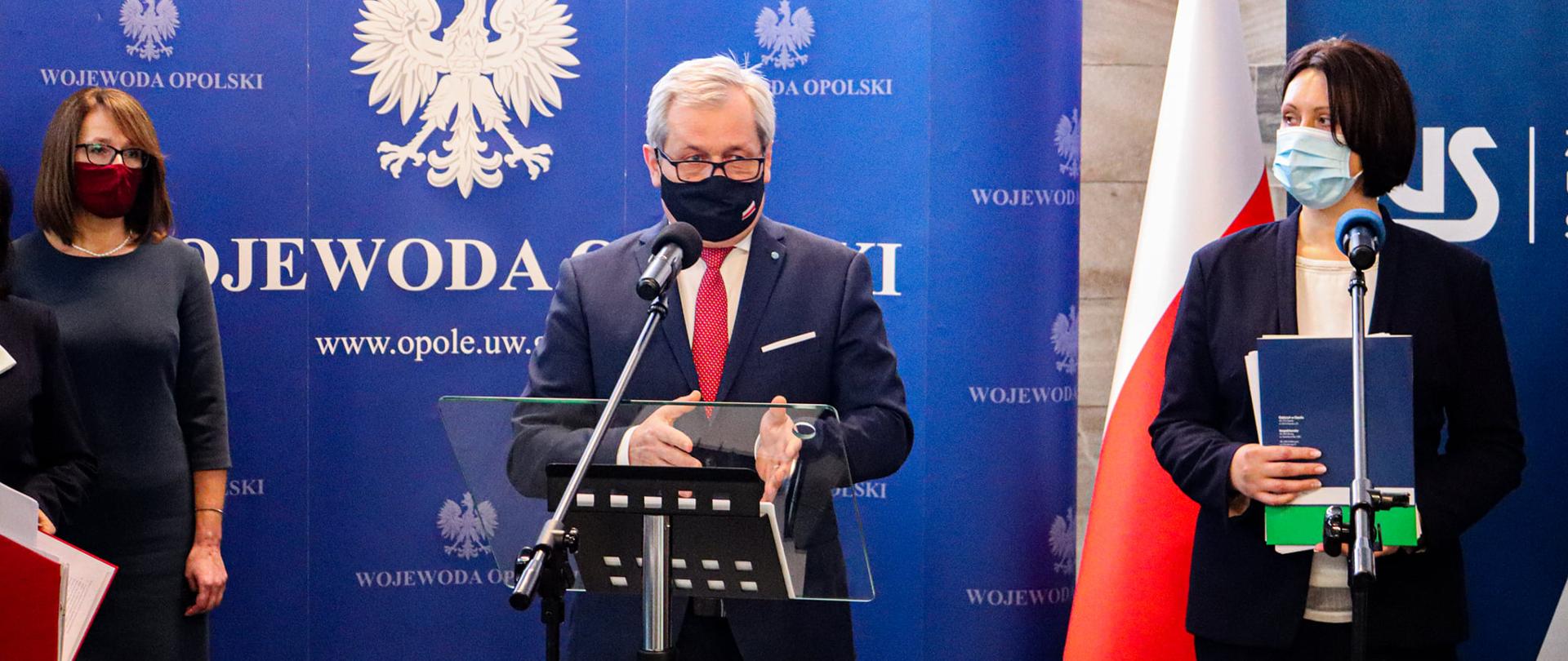 Na zdjęciu wojewoda opolski, przemawiający zza mikrofonu na statywie. Po jego lewej stronie dyrektorka opolskiego ZUS, po prawej dyrektorka KAS w Opolu. W tle ścianka z logotypem Wojewody Opolskiego.