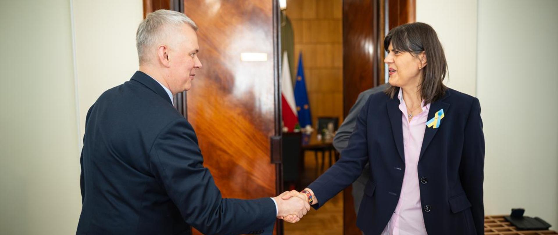 Na zdjęciu witający się ze sobą minister Tomasz Siemoniak (po lewej) oraz europejska prokurator generalna Laura Codruta Kövesi (po prawej). W tle widać brązowe drzwi i fragment wnętrza sali (w środku: stolik, krzesła, flagi Polski i UE). Po prawej stronie zdjęcia widać drewniany mebel.