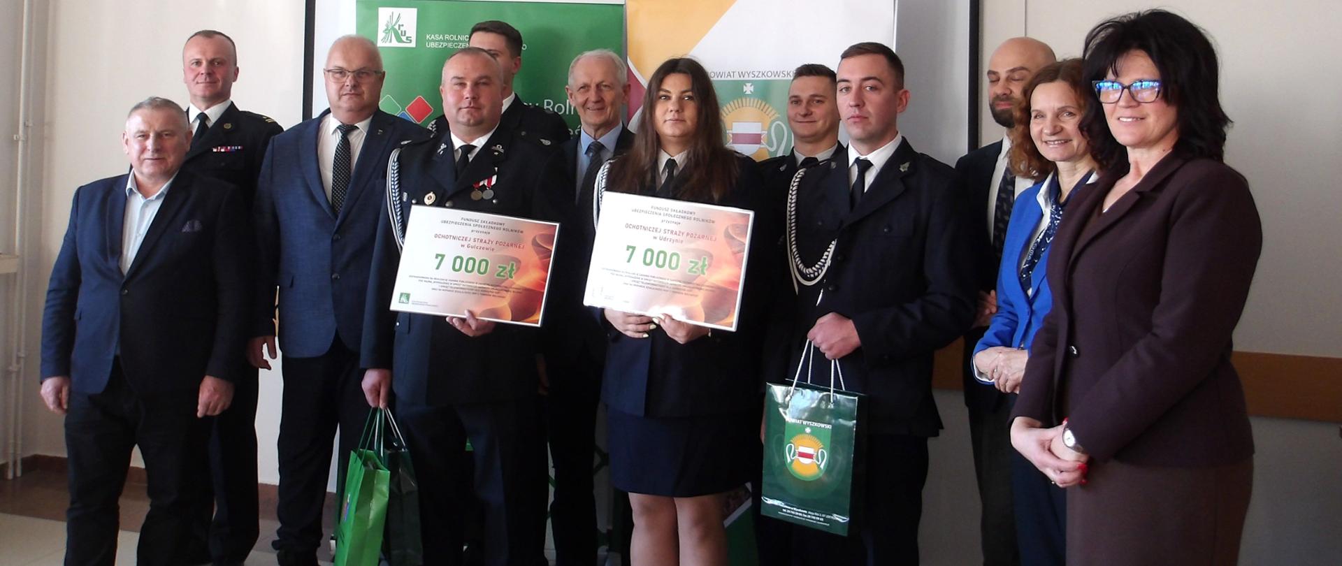 Dziewięciu mężczyzn i trzy kobiety stoją na tle banerów KRUS i Powiatu Wyszkowskiego. Kobieta i mężczyzna w galowych mundurach strażaka trzymają planszę z wysokością promesy siedmiu tysięcy złotych.