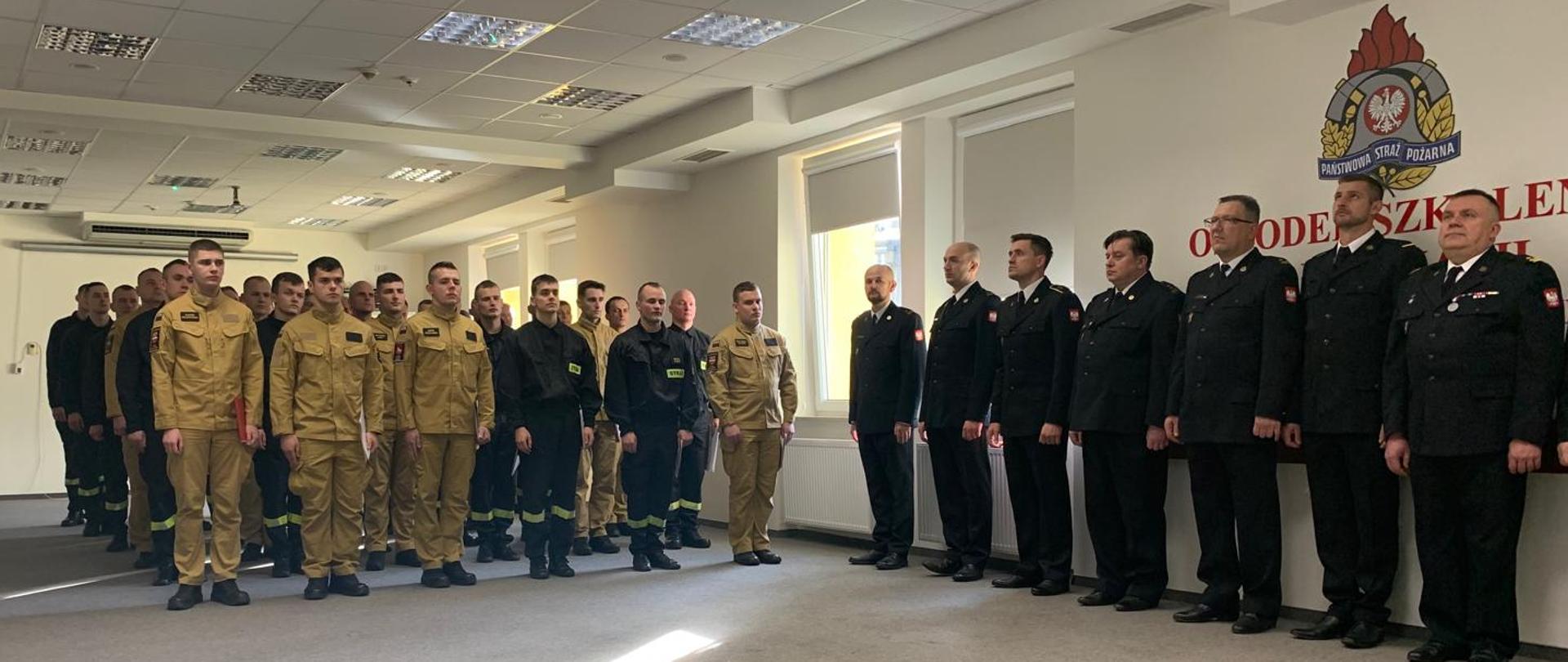 Zastępca Mazowieckiego Komendanta Wojewódzkiego PSP bryg. Waldemar Wysowski i dwóch strażaków PSP stoją po prawej stronie zdjęcia. Po lewej stronie pod ścianą stoją inni strażacy i pracownicy. W pomieszczeniu są beżowe ściany i szaro-beżowa wykładzina. Przez okno widoczne są drzewa.