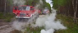 Na zdjęciu widać leśną drogę z ustawionymi samochodami pożarniczymi, duże pozorowane zadymienie.