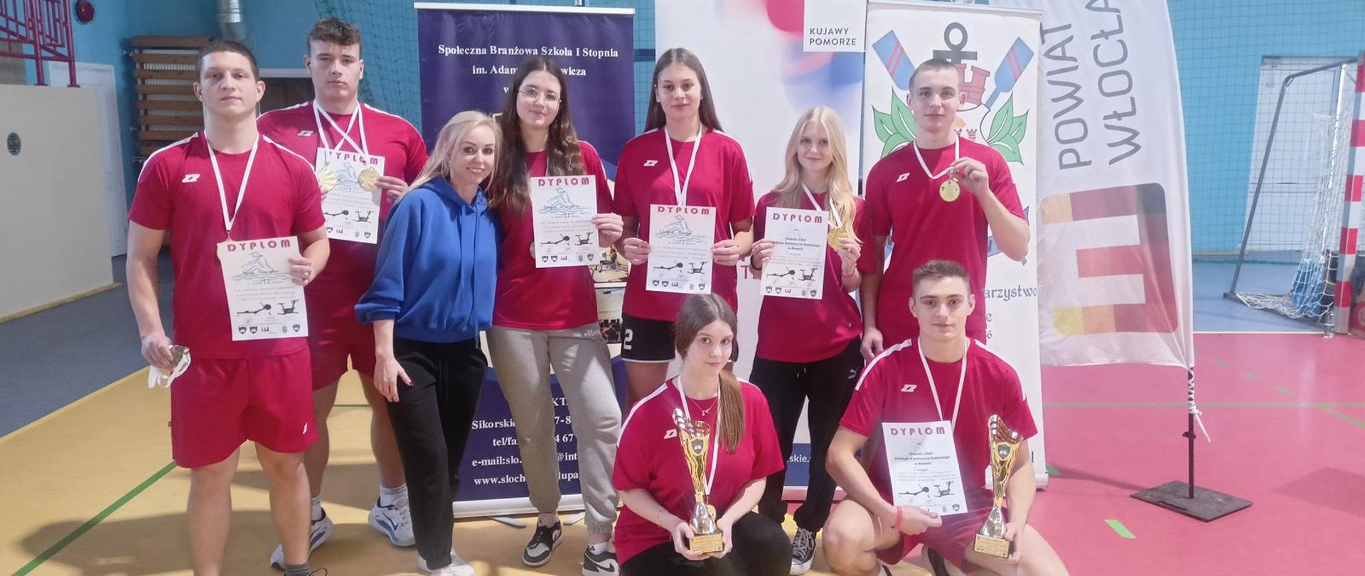 Na zdjęciu grupa uczniów Zespołu Szkół CKR w Kowalu pozuje w sali gimnastycznej po zakończonych zawodach sportowych. Wszyscy trzymają w dłoniach dyplomy, medale i złote puchary. W środku grupy stoi nauczycielka wychowania fizycznego w niebieskiej bluzie z uśmiechem obejmująca uczniów. Uczniowie prezentują radość i dumę ze zdobytych nagród